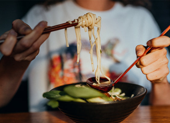 Frauenhände halten Essstäbchen in der einen und Suppenlöffel in der anderen Hand und zeigen die fertigen Ramennudeln